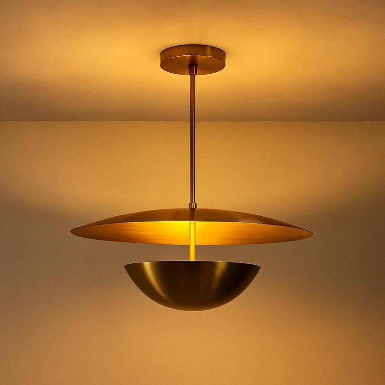 May include: A modern, brass-toned pendant light fixture. The design features a large, flat disc above a bowl-shaped shade, suspended from a ceiling mount by a slender rod. The warm gold color suggests an elegant, contemporary aesthetic.