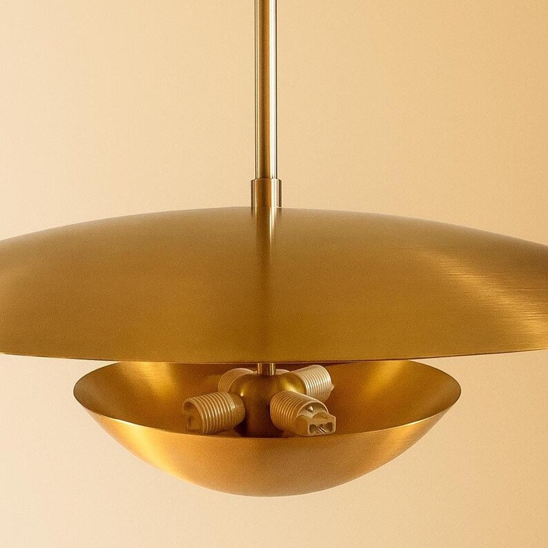 May include: A gold-colored, mid-century modern pendant light fixture. The fixture has a large, flat, circular shade above a bowl-shaped light housing. The light housing contains three light bulb sockets.
