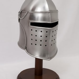 May include: A silver-colored medieval helmet with a pointed top, displayed on a wooden stand. The helmet features a visor with a horizontal slit and numerous small ventilation holes. A decorative chain is attached to the side.