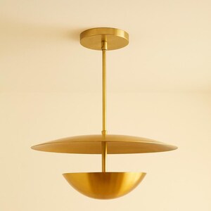 May include: A modern, gold-colored pendant light fixture. The fixture has a circular ceiling mount, a long, straight rod, a large, flat, disc-shaped shade, and a bowl-shaped bottom. The light is suspended from the ceiling.