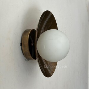 May include: A wall-mounted light fitting with a brass finish and a white globe-shaped light. The fitting has a unique design with a curved, leaf-like shape surrounding the light. The text "Lighting Space" is visible.