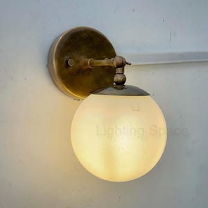 May include: A wall-mounted light fitting featuring a round, frosted glass globe. The fixture has a brass-coloured metal base and arm. The text "Lighting Space" is visible on the globe.