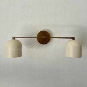 May include: A wall-mounted light fixture with two off-white, dome-shaped shades. The fixture has a bronze-colored circular base and a horizontal arm. The text "Lighting Space" is visible on the left shade.