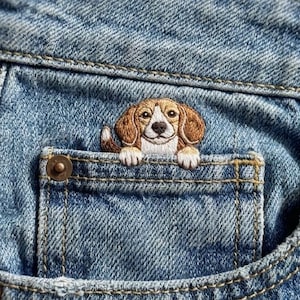 Può includere: Jeans in denim azzurro con un piccolo e dettagliato ricamo di un cane beagle marrone e bianco che spunta dalla parte superiore della tasca portamonete anteriore. Il ricamo mostra la testa del cane e le zampe anteriori sul bordo.