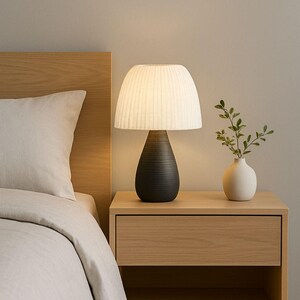 May include: A modern bedroom scene featuring a bedside table with a lamp and a small vase. The lamp has a white, pleated shade and a dark, textured base. A small white vase holds a sprig of greenery.