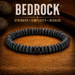 May include: A circular bracelet composed of numerous small, dark gray, disc-shaped beads and one smooth, matte black spherical bead. The text BEDROCK, STRENGTH, SIMPLICITY, RESOLVE is displayed in white and gold lettering above the bracelet.