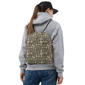 May include: A drawstring backpack with a repeating geometric pattern in shades of brown and beige. The bag is worn over the shoulders with black straps. The person is wearing a gray hoodie, blue jeans, and a black baseball cap.