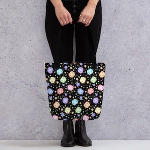 May include: Black tote bag featuring a repeating pattern of pastel dice, stars, and leaves. The bag is held by a person wearing black trousers and boots. The bag has black handles.
