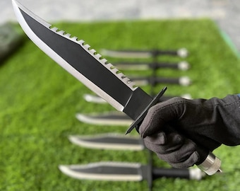May include: A black and silver survival knife with a serrated edge is held in a gloved hand. Several similar knives are arranged on a green surface in the background. The knife has a black handle and a silver blade.