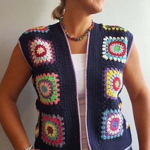 May include: A navy blue crochet waistcoat with a purple trim and colourful granny square designs. The waistcoat is sleeveless and has a V-neck.