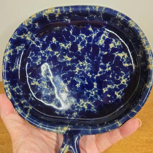 Pode incluir: Um prato de cerâmica azul com uma alça, apresentando um padrão salpicado de amarelo e branco sobre um fundo azul escuro. O prato tem formato arredondado e acabamento brilhante, adequado para servir ou expor.