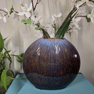 May include: A spherical ceramic vase with a blue and brown striped design. The vase contains white flowering branches. The vase is placed on a light blue surface, with a green plant visible in the background. The vase is a decorative home decor item.