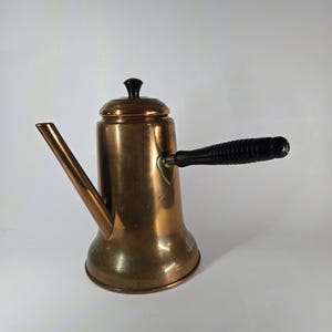 Può includere: Una caffettiera color rame con manico e pomello del coperchio neri. La caffettiera ha un lungo beccuccio e una base arrotondata. Il manico è in legno nero. La caffettiera è probabilmente vintage e in buone condizioni.