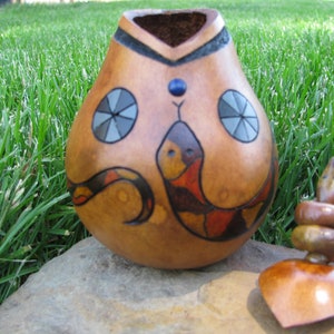 May include: A hand-painted gourd with a snake design. The gourd is brown with red, orange, and black accents. The snake is curled around the gourd and has a blue eye. The gourd is sitting on a rock.