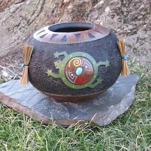 May include: A hand-painted gourd with a black, brown, green, and turquoise design. The gourd is decorated with a stylized lizard and a circular pattern. The gourd is sitting on a slate stone.