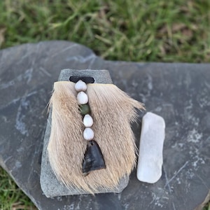 May include: A handcrafted pendant featuring a rectangular stone base with a fur accent. The pendant is adorned with white and green beads, and a dark, triangular charm. A white stone rests beside the pendant, set against a gray stone surface.