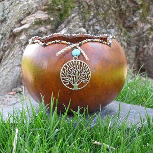 May include: A brown gourd with a woven rim and a metal tree of life pendant hanging from it. The gourd is sitting on a stone surface with green grass in the background.