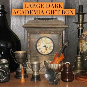 May include: A large dark wooden box with a clock face and a small violin inside. The box is surrounded by various items, including a globe, a candlestick, a glass bottle, a camera, and several trophies. The text "LARGE DARK ACADEMIA GIFT BOX" is displayed above the box. The text "PRISM ARTWORKS & FURNITURE" is displayed in a green rectangle at the bottom right corner of the image.