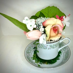 Op de afbeelding: Een wit theekopje met een bloemenpatroon is gevuld met een boeket roze en witte bloemen. De bloemen omvatten rozen, tulpen en baby's adem. Het theekopje staat op een bijpassend schoteltje.