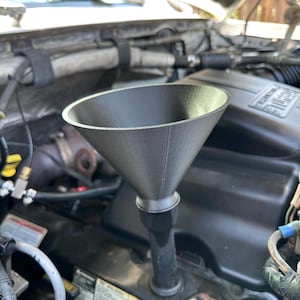 May include: A gray funnel with a wide mouth and a narrow spout is positioned over a car engine. The funnel is made of a matte material and is designed to aid in the pouring of fluids into the engine.