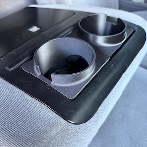 May include: A black plastic automotive center console insert featuring two circular cup holders with side cutouts, integrated into a light gray fabric seat surface. The console includes a flat, rectangular storage compartment lid adjacent to the cup holders.