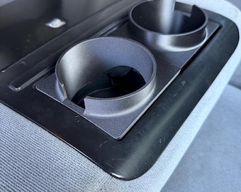 May include: A black plastic automotive center console insert featuring two circular cup holders with side cutouts, integrated into a light gray fabric seat surface. The console includes a flat, rectangular storage compartment lid adjacent to the cup holders.