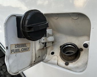 Op de afbeelding: Open brandstofklep van een wit voertuig, met een zwarte tankdop en de vulhals. De deur heeft een sticker met de tekst "DIESEL FUEL ONLY". De brandstofklep is open.