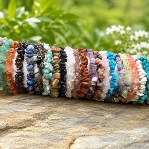 Könnte beinhalten: Eine Sammlung mehrerer Stretch-Armbänder aus verschiedenen polierten Edelstein-Splittern in einer Vielzahl von Farben, darunter Weiß, Grün, Orange, Schwarz, Blau, Braun, Violett, Rosa, Türkis und Gelb, die in einer horizontalen Reihe angeordnet sind.
