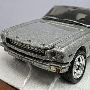 May include: Silver convertible toy car with black tyres and chrome rims. The model car has a detailed front grille and headlights. The car's interior is black, and the top is white. The car has a Ford logo on the front.