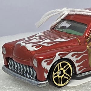 May include: A red and gold toy car with white flame decals. The car has a wood-paneled side and gold-colored wheels. A white string is attached to the top of the car, suggesting it is an ornament.
