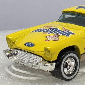 Puede incluir: Un coche de juguete amarillo con el logotipo de Nesquik y el número 10 en el lateral. El coche tiene neumáticos de banda blanca y un parachoques cromado. El logotipo de Ford es visible en la parte delantera.
