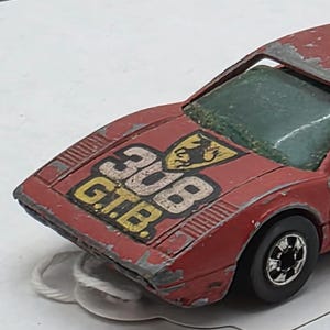May include: A vintage red die-cast toy car with the text "308 G.T.B." on the hood. The car has black wheels and a weathered appearance, suggesting it is a collectible item. The car's length is approximately 5 inches.
