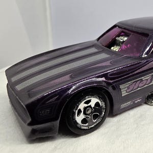 May include: A purple Hot Wheels toy car with silver accents. The car features a sleek design with a spoiler and the words "Hot Wheels" on the side. The hood has three silver stripes. The windows are tinted purple.