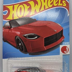 May include: A red Hot Wheels die-cast car, model 3/10, in a clear plastic package. The car is a sleek sports car design with black wheels and a black roof. The packaging has the Hot Wheels logo and the text "HW J-Imports".