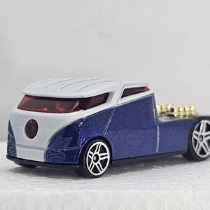 May include: A die-cast toy truck with a white cab and a blue body. The truck features a unique design with a low profile and custom wheels. The rear bed has gold-colored engine detailing. The vehicle is a collectible toy.