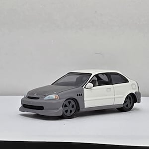 May include: A two-door model car, painted in white and gray, with black wheels. The car has a sporty design, with a low profile and a spoiler. The car is a miniature replica of a real vehicle.