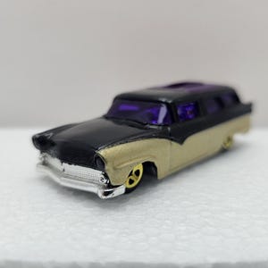 May include: A die-cast model of a classic station wagon. The car is painted black on top and gold on the bottom, with purple tinted windows. The wheels are gold and the front bumper is silver.