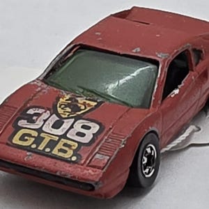 May include: A vintage red toy car with the text "308 G.T.B." on the hood. The car has black wheels and a green windshield. The paint shows signs of wear, indicating it's a well-loved collectible.
