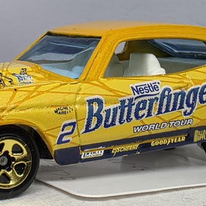 May include: A yellow die-cast toy car with gold-coloured wheels and black tyres. The car features the text "Nestle Butterfinger World Tour" and the number "2" on the side. The car has a silver front bumper and a white interior.