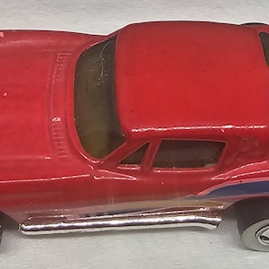 May include: A red toy car with a sleek design, featuring a low profile and a curved roof. The car has silver side pipes, black tires, and white rims. Blue and yellow stripes accent the side.