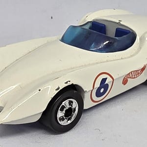 Può includere: Un'auto da corsa giocattolo bianca vintage con parabrezza e interni blu. L'auto presenta ruote nere e il numero 6 in un cerchio blu con bordo rosso. È visibile un logo Hot Wheels.