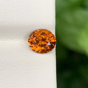 Könnte beinhalten: Ein runder, facettierter, orangefarbener Edelstein. Der Edelstein liegt vor einem weißen Hintergrund, mit einem verschwommenen grünen Hintergrund. Der Edelstein hat einen Brillantschliff, der das Licht reflektiert und seine Farbe zeigt.