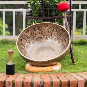 以下が含まれることがあります： 瞑想中の人物の複雑なエッチングデザインが施された、大きく装飾されたシンギングボウル。赤いフェルトの頭が付いた木製のマレットが近くにあり、小さな木製と黒色のマレットもあります。ボウルは丸いタン色のクッションの上にあります。