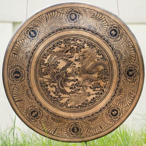 Peut inclure: Un grand gong rond de couleur bronze avec des gravures complexes. Le motif central repr&eacute;sente un dragon et un ph&eacute;nix parmi des nuages. Le bord ext&eacute;rieur pr&eacute;sente des motifs floraux et des symboles. Le gong est suspendu par une fine corde, avec de l'herbe verte au premier plan.