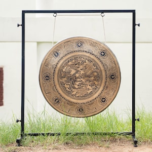 Peut inclure: Un grand gong orn&eacute; d'un motif de dragon d&eacute;taill&eacute;, suspendu &agrave; un cadre en m&eacute;tal noir. Le gong est de couleur or chaud avec des gravures complexes. Le cadre est pos&eacute; sur une zone herbeuse, avec un mur blanc en arri&egrave;re-plan.