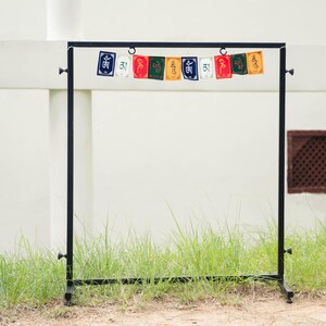 Peut inclure: Un pr&eacute;sentoir en m&eacute;tal noir avec des drapeaux de pri&egrave;re color&eacute;s suspendus en haut. Les drapeaux sont rectangulaires et pr&eacute;sentent diverses couleurs et symboles. Le pr&eacute;sentoir se dresse sur de l'herbe devant un mur blanc.