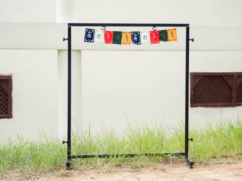 Peut inclure: Un pr&eacute;sentoir en m&eacute;tal noir avec des drapeaux de pri&egrave;re color&eacute;s suspendus en haut. Les drapeaux sont rectangulaires et pr&eacute;sentent diverses couleurs et symboles. Le pr&eacute;sentoir se dresse sur de l'herbe devant un mur blanc.