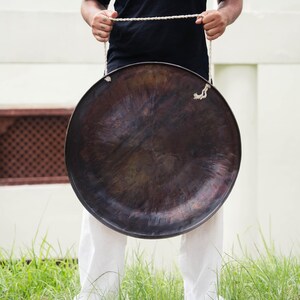 Peut inclure: Un grand gong en bronze fonc&eacute; suspendu par une corde blanche. Le gong est tenu devant une personne portant un t-shirt noir et un pantalon blanc. L'arri&egrave;re-plan comprend un mur blanc et de l'herbe verte.