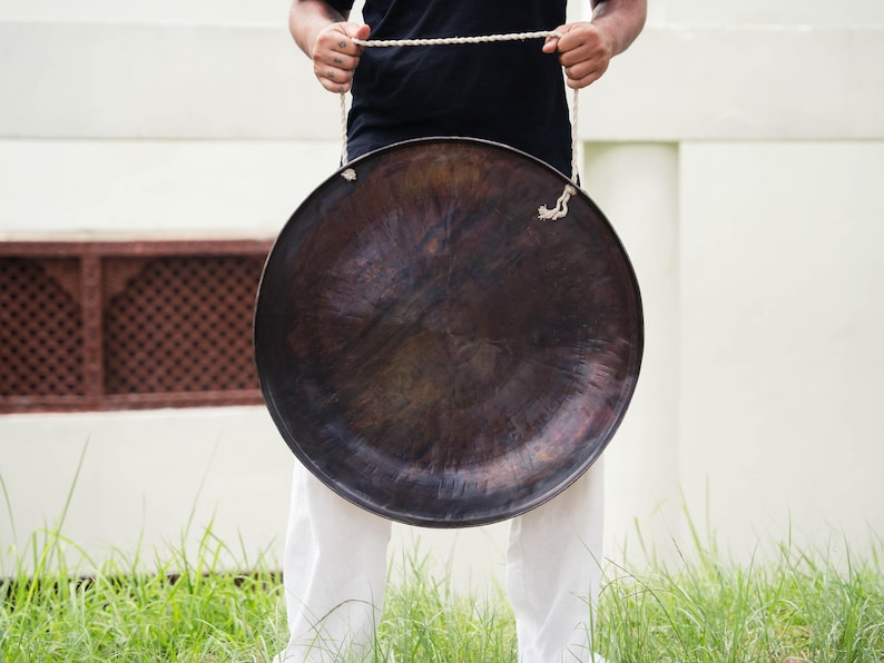 Peut inclure: Un grand gong en bronze fonc&eacute; suspendu par une corde blanche. Le gong est tenu devant une personne portant un t-shirt noir et un pantalon blanc. L'arri&egrave;re-plan comprend un mur blanc et de l'herbe verte.