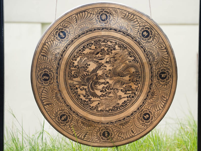 Peut inclure: Un grand gong rond de couleur bronze avec des gravures complexes. Le motif central repr&eacute;sente un dragon et un ph&eacute;nix parmi des nuages. Le bord ext&eacute;rieur pr&eacute;sente des motifs floraux et des symboles. Le gong est suspendu par une fine corde, avec de l'herbe verte au premier plan.